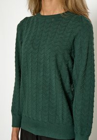 Suéter verde de punto con un patrón de chevron texturizado, cuello redondo y mangas largas con puños y dobladillo acanalados.