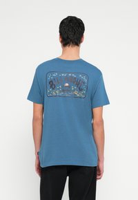 Blå bomulds-T-shirt med et retro grafisk tryk på ryggen, med palmer og bølger. Korte ærmer og en rund halsudskæring.