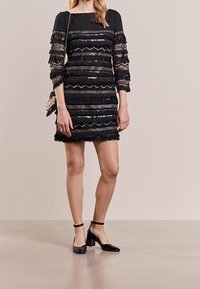 Robe noire avec des sequins, des franges et des rayures texturées. Manches trois-quarts et décolleté carré. Associée à des chaussures à talons carrés noires.