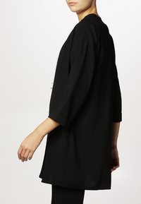 Černý oversized cardigan s tříčtvrtečními rukávy, jemnou texturou a uvolněným střihem, s minimálním designem a bez viditelných zapínání.