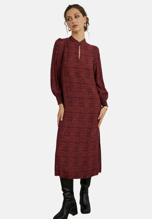 Mujer con vestido midi de manga larga color burdeos con estampado, escote con abertura y botas negras hasta la rodilla, posando frente a un fondo liso.