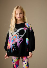 Sweatshirt noir oversize avec graffiti de cœur coloré, associé à des leggings vibrants ornés de motifs et formes abstraites.