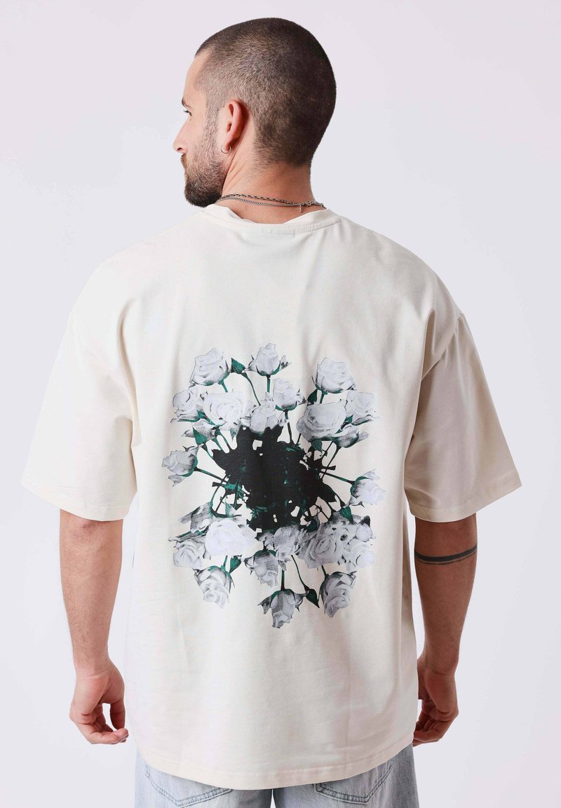 Homme aux cheveux courts portant un t-shirt blanc surdimensionné avec un motif circulaire floral noir et blanc dans le dos, vu de profil.