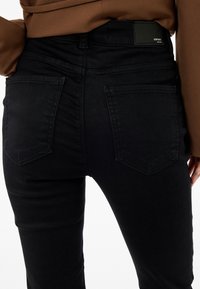 Jeans de mezclilla negra con un diseño ajustado, que cuentan con bolsillos traseros y una textura suave. La etiqueta negra en la cintura indica la marca.