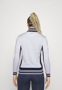 Hvid atletisk jakke med en marineblå stribet krave og kant, tætsiddende design, lange ærmer og en glat tekstur. Logo vist på ryggen.