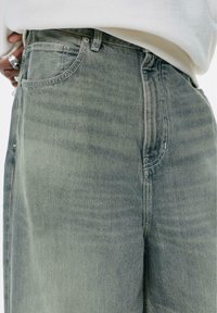 Jeanși din denim verde deschis au un design clasic cu cinci buzunare, o textură decolorată și detalii de cusătură vizibile la cusături și buzunare.