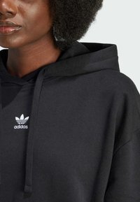 Svart hettegenser med strukturert stoff, snortrukket hette og en hvit Adidas-logo på brystet. Myk og avslappet design.