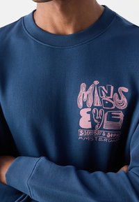 Granatowa bluza z miękkiego materiału z różowym nadrukiem "MINDS EYE" oraz "SCOTCH & SODA AMSTERDAM" na przodzie.