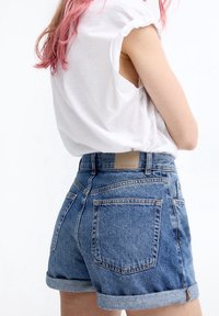 Denim shorts with rolled cuffs, light blue wash, and a tan leather patch on the back. Paired with a white short-sleeve shirt.