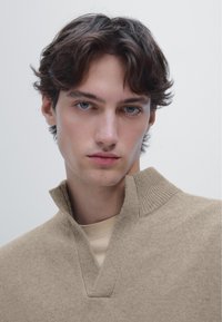 Beige strikket pullover med høj krave og en smal V-hals. Blød tekstur, tætsiddende design, båret over en lys beige T-shirt, minimalistisk stil.