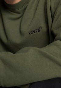 Personne portant un sweat-shirt Levi's vert olive avec un logo noir brodé sur la poitrine, les bras croisés reposant sur une surface.