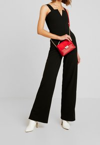 Combinaison noire à jambes larges, décolleté en V profond et bretelles épaisses. Associée à un petit sac rouge avec des clous et une bandoulière en chaîne. Talons blancs.