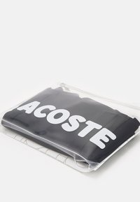 Vêtement noir emballé dans une pochette zippée en plastique transparent, avec un grand logo blanc "LACOSTE", d'apparence plate et texturée.