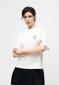 Άτομο με κοντά σκούρα μαλλιά στέκεται με τα χέρια σταυρωμένα, φορώντας ένα λευκό T-shirt Carhartt WIP και μαύρη φούστα μπροστά σε απλό φόντο.