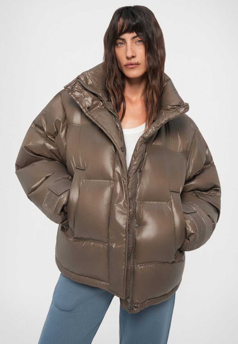 Vrouw met lang donker haar, gekleed in een oversized glanzende bruine pufferjack, witte top en blauwe broek tegen een effen achtergrond.