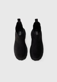Une paire de bottines noires en daim avec des tirettes et des semelles épaisses en caoutchouc, vues de dessus sur un fond blanc.