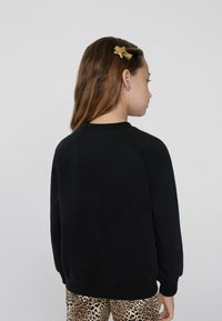 Černá mikina s raglánovými rukávy, strukturovaná tkanina a žebrované manžety, nošená přes béžové kalhoty s leopardím potiskem. Vlasy upravené zlatou hvězdičkou ve vlasech.