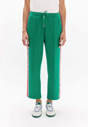 Persona con pantalones verdes con cordón, con rayas laterales rojas y blancas y zapatillas blancas con detalles azules y verdes.