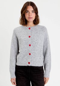 Femme aux cheveux bruns mi-longs portant un pull en laine moelleux gris clair avec des boutons en forme de cœur rouge et un pantalon noir, se tenant droit.