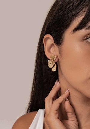 Goldene Ohrringe in Form von stilisierten Blättern mit einer strukturierten Oberfläche. Das Modell hat dunkles Haar und trägt ein weißes Oberteil.
