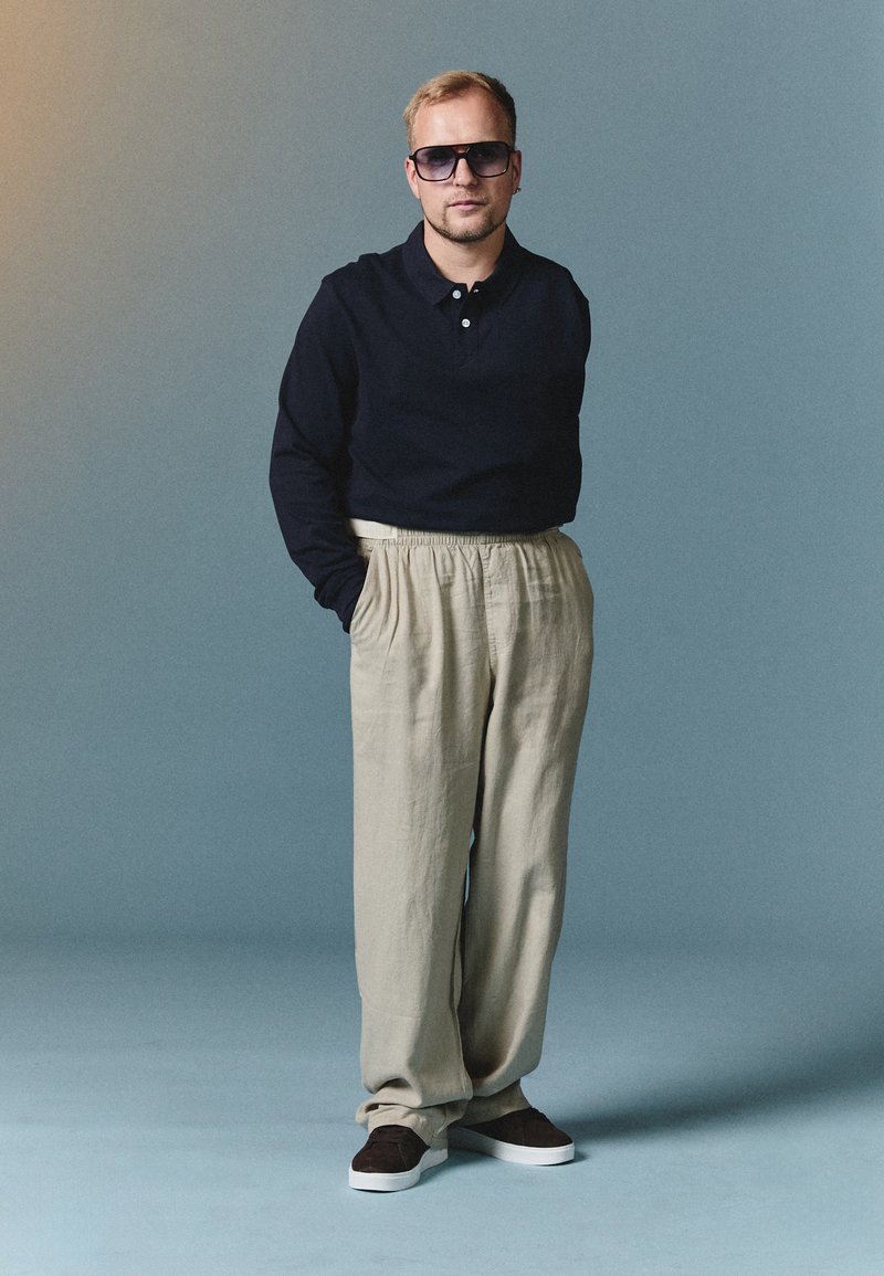 Polo albastru cu mâneci lungi, pantaloni din in bej, ochelari de soare negri și adidași maro. Modelul stă cu mâinile în buzunare, pe un fundal albastru.