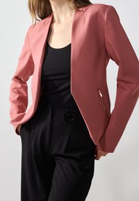 Rosa skräddarsydd blazer med en slank design, som har en subtil glans, öppen framsida och dragkedjefickor på sidan. Matchad med svarta hög midja byxor.