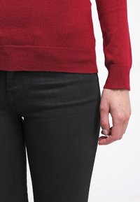 Gros plan d'une personne portant un pull rouge à manches longues et un pantalon noir, montrant le torse et la main droite reposant le long du corps.