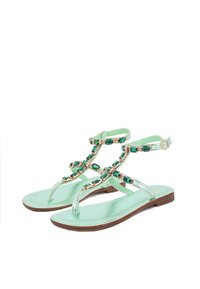 Sandali piatti verde menta con finitura lucida, caratterizzati da dettagli in tono oro e strass ematiti verdi, con cinturini alla caviglia regolabili e una suola marrone.