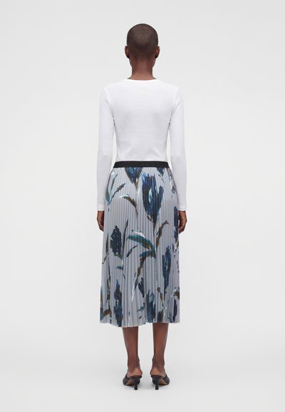 Femme aux cheveux courts portant un haut blanc à manches longues, une jupe midi plissée à imprimé floral bleu et des chaussures à talons noires, vue de dos.
