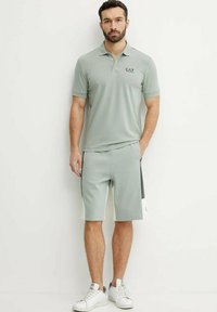 Polo shirt in poliestere grigio chiaro con maniche corte, abbinato a pantaloni corti coordinati. Presenta un logo nero, dettagli bianchi e un design atletico.