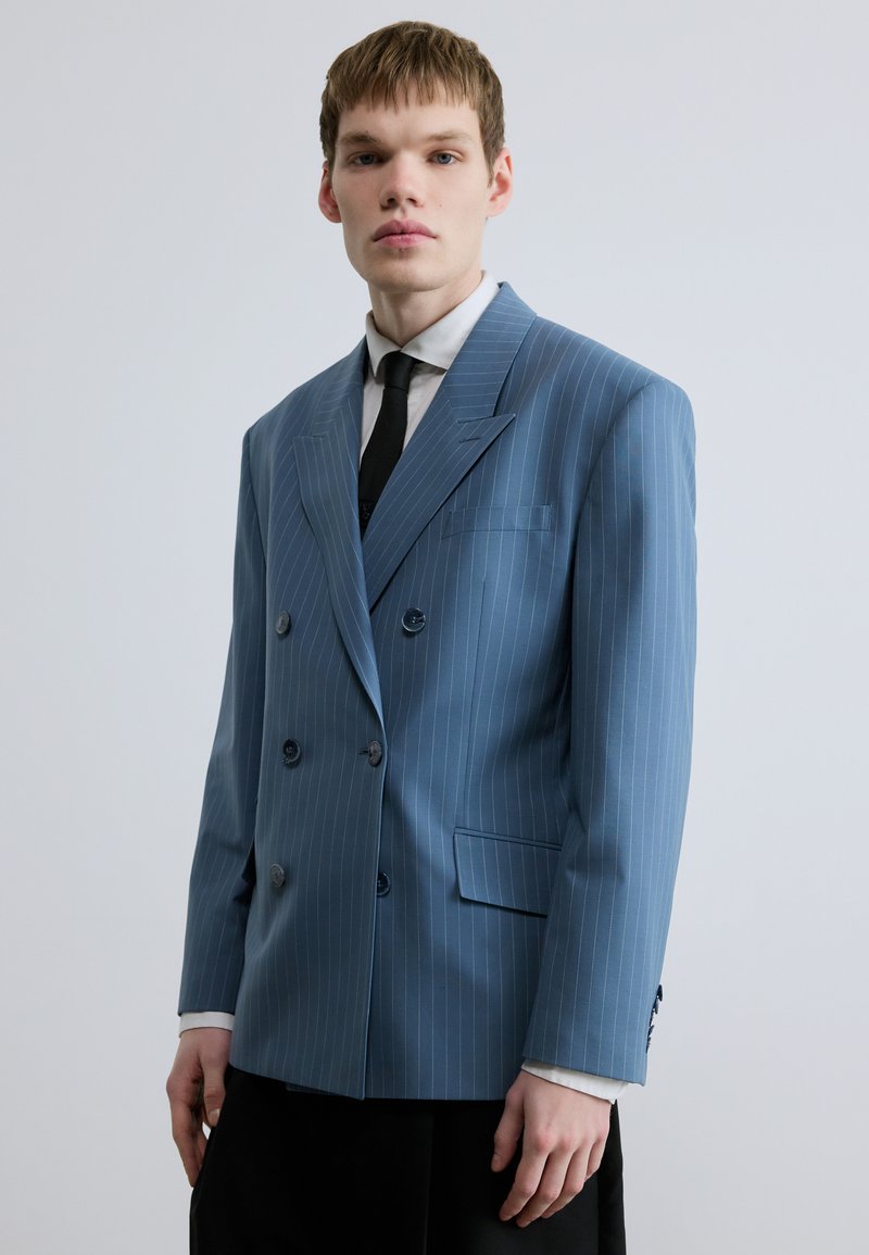 Sacou dublu cu dungă albastră, rever decupat, doi nasturi, un buzunar la piept și material texturat. Cravată neagră purtată dedesubt.