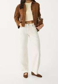 Woman wearing a brown cropped jacket, cream crop top, white high-waisted pants, brown flats, and holding a studded brown clutch.