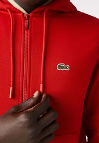 Närbild av en mörkhyad hand som justerar den röda dragkedjan på en röd Lacoste-hoodie med en grön krokodillogotyp på bröstet.