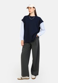 Marine-blaues Oversized-T-Shirt über einem hellblauen, gestreiften Hemd, kombiniert mit grauen, weit geschnittenen Hosen und beigen Sneakers. Weiße Kappe. Silberne Halskette.