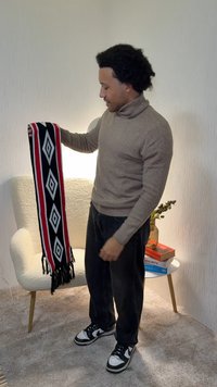 Stickad svart halsduk med röda och vita geometriska mönster, fransiga kanter, hålls av en person som bär en beige polotröja och mörka jeans.