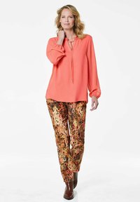 Korallefarbige plissierte Bluse mit einem Bindekragen und langen Ärmeln, kombiniert mit gemusterten goldenen und blumigen Hosen. Braune Stiefeletten runden das Outfit ab.