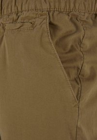 Khaki shorts med en glat tekstur, der har en elastisk talje og detaljer med sidelommer, vist fra en tæt vinkel.