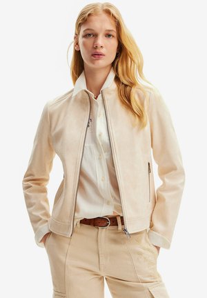 Giacca in suede beige con zip frontale, colletto alla cinese e tasche laterali, indossata sopra una camicia bianca con bottoni e pantaloni beige.