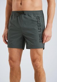 Costumi da bagno verde scuro con vita elasticizzata, dettagli con cordoncino, tasche laterali e logo verticale nero sulla gamba sinistra.