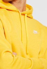 Sudadera amarilla de Nike con tela de mezcla de algodón, que incluye un bolsillo canguro frontal, cordones ajustables y un logo bordado en blanco en el pecho.