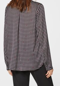 Kvinna som bär en långärmad svart blus med ett mönster av små vita och rosa rutor, tillsammans med svarta byxor, sedd bakifrån.