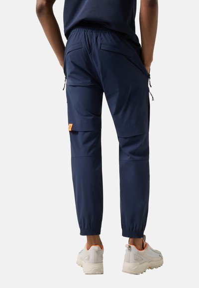 Osoba ubrana w granatowe spodnie typu cargo z elastycznymi ściągaczami u dołu oraz białe trampki, stojąca tyłem z rękami w kieszeniach.