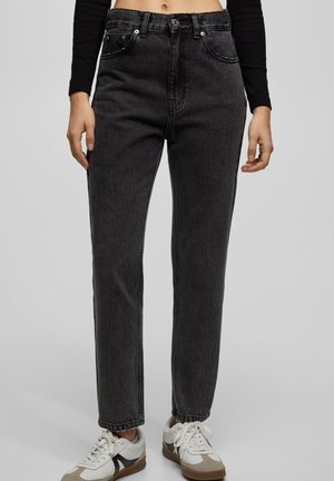 Femme portant un jean noir taille haute associé à des baskets blanches, debout devant un fond gris uni.