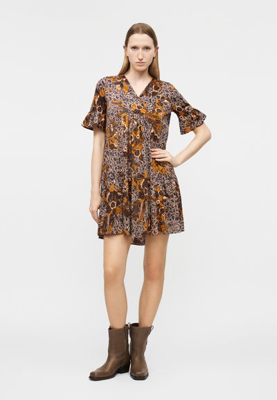 Γυναίκα στέκεται με το χέρι στη μέση φορώντας κοντό φόρεμα με λουλουδάτο μοτίβο και βολάν στα μανίκια, καθώς και καφέ μποτάκια αστραγάλου, μπροστά σε απλό ανοιχτόχρωμο φόντο.