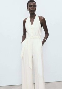 Jumpsuit couleur crème avec un col halter, des boutons avant, une taille ceinturée et des jambes larges, présentant un tissu texturé et des poches latérales.