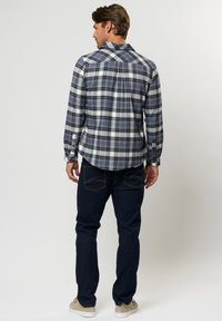 Mees seisab seljaga vaataja poole, kandes sinist, valget ja pruuni ruudulist särki, tumedaid teksaseid ja beeže tossusid ühetaustalise heleda tausta ees.