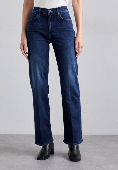 Jean bleu foncé en denim, à la coupe droite, taille haute, et avec des coutures visibles. Porté avec des bottines noires.