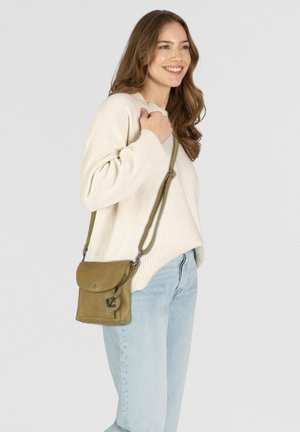 Donna sorridente che indossa un maglione crema e jeans blu chiaro, mentre porta una borsa a tracolla verde oliva con un emblema ad ancora.