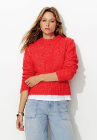Pull en maille rouge avec un motif texturé, col rond et poignets côtelés. Porté sur une chemise blanche, associé à un jean en denim bleu clair.