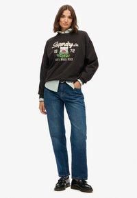 Czarna bluza z logo "Superdry.ca", zestawiona z jasnoniebieską koszulą w paski pod spodem oraz niebieskimi dżinsami prostymi. Noszona z czarnymi butami.